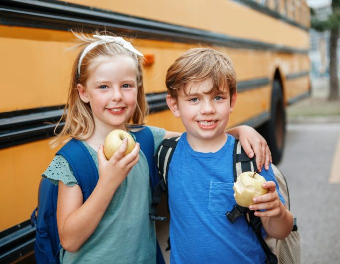 Gelber Schulbus fährt auf einer Straße entlang, im Hintergrund Häuser und grüne Bäume.